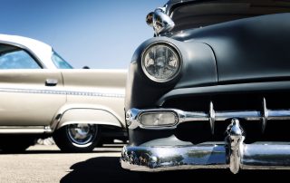 Närbild av framändarna på två klassiska bilar parkerade sida vid sida, med vintagedesigner med kromdetaljer under en klarblå himmel.